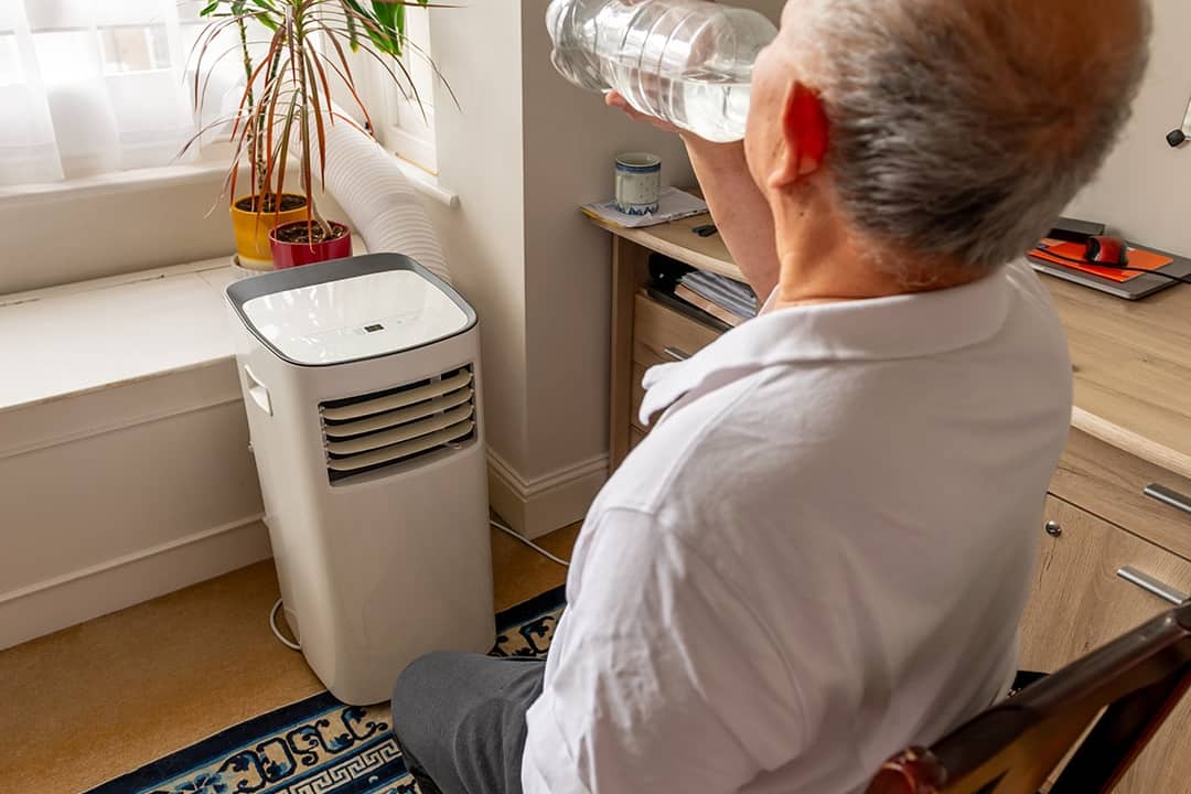An elderly man drinking water and cooling himself in front of an AC unit during summer.